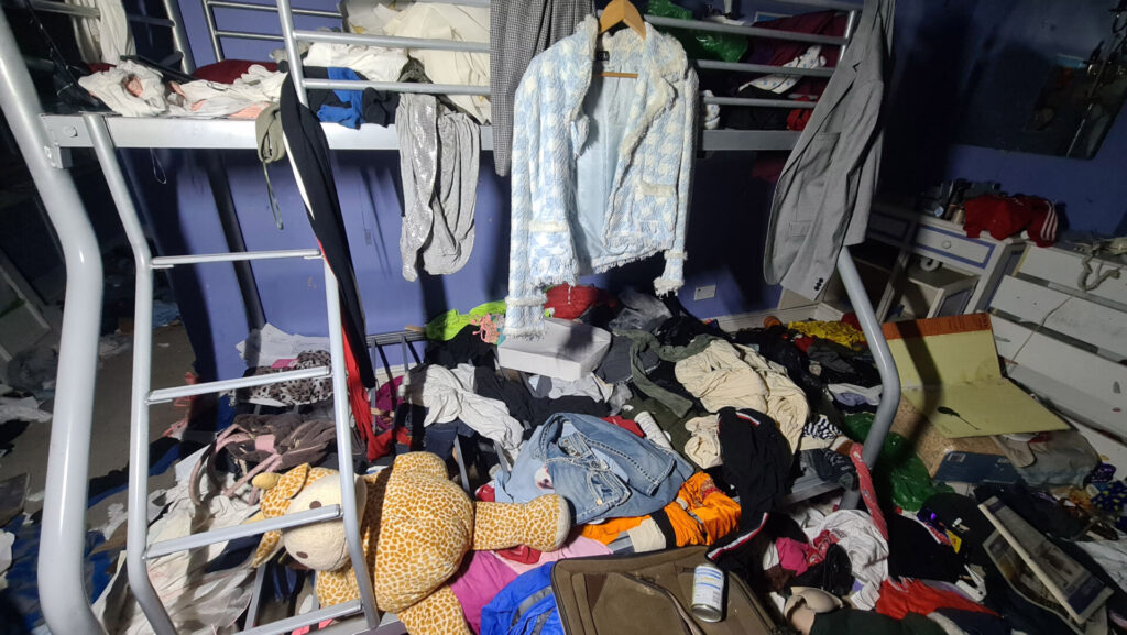 A blue checkered jacket and other clothes, as well as toys, are thrown on the floor and hang from the metal bunk beds in the purple room. 
