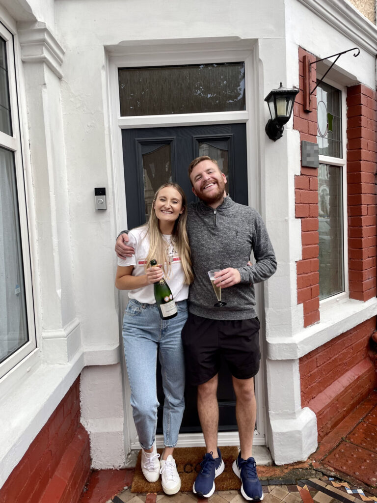 Elle & Alex smiling outside their home with a bottle of fizz
