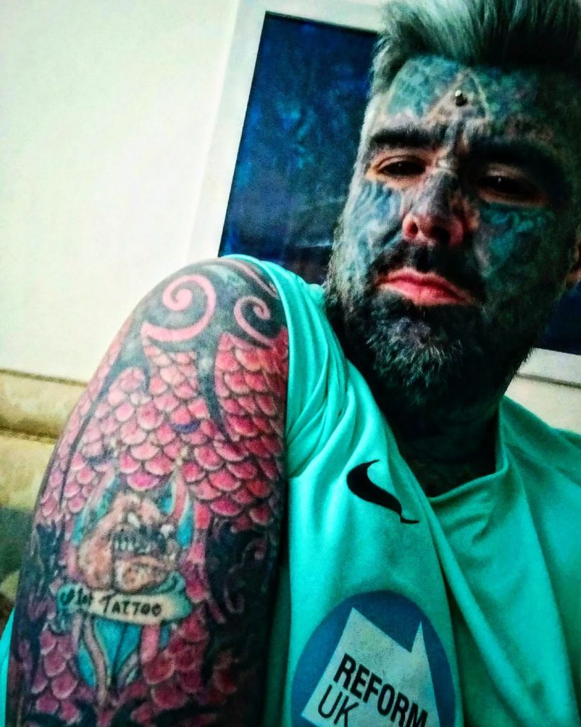 King Of Ink Land poses for a selfie while wearing a green top and showing his bulldog tattoo on his arm. 