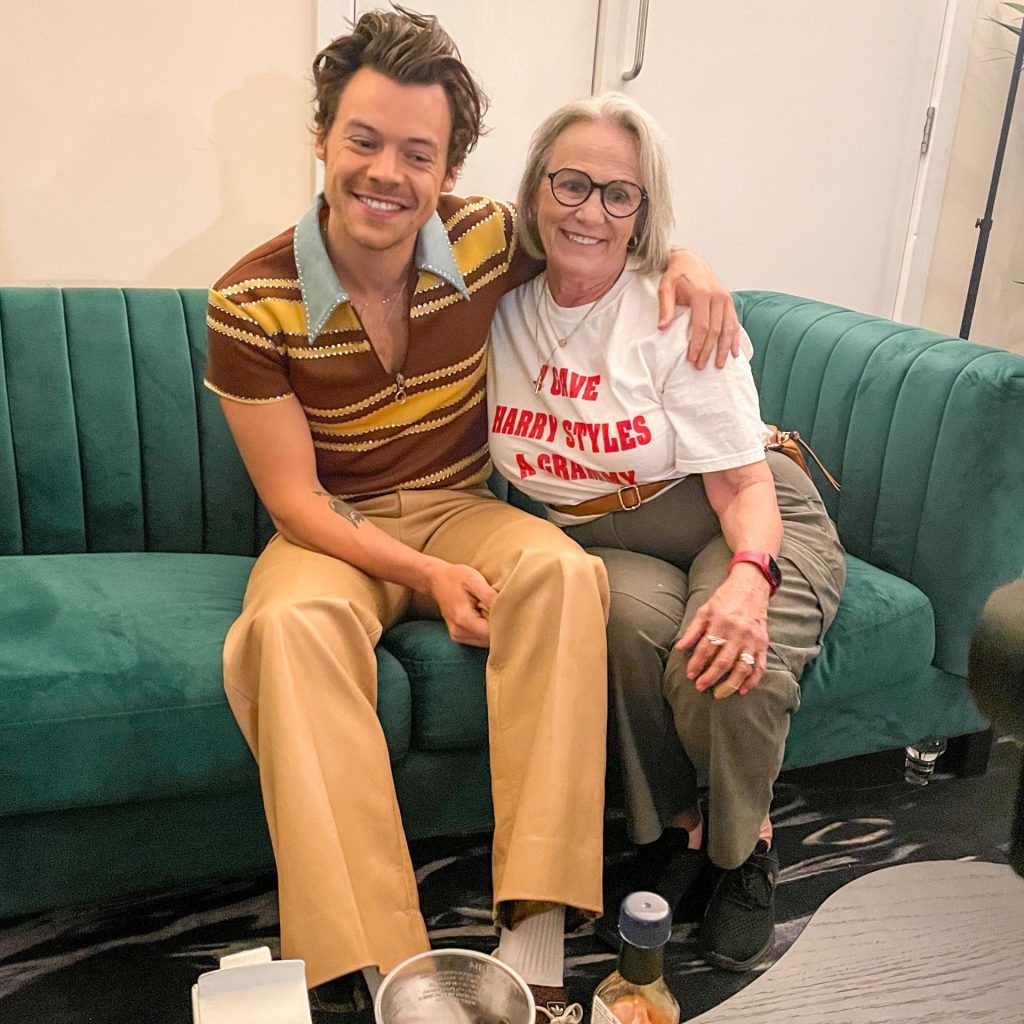 Reina smiles while sitting on a green sofa next to Harry Styles who is smiling and has his arm around her.