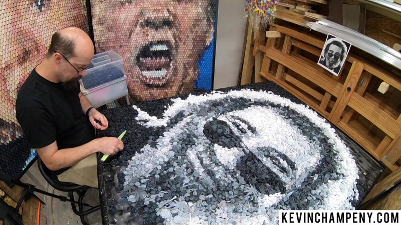 Kevin is shown working on a piece which is black and white of a man's face with a beard and sunglasses, with the picture in the background for reference. 