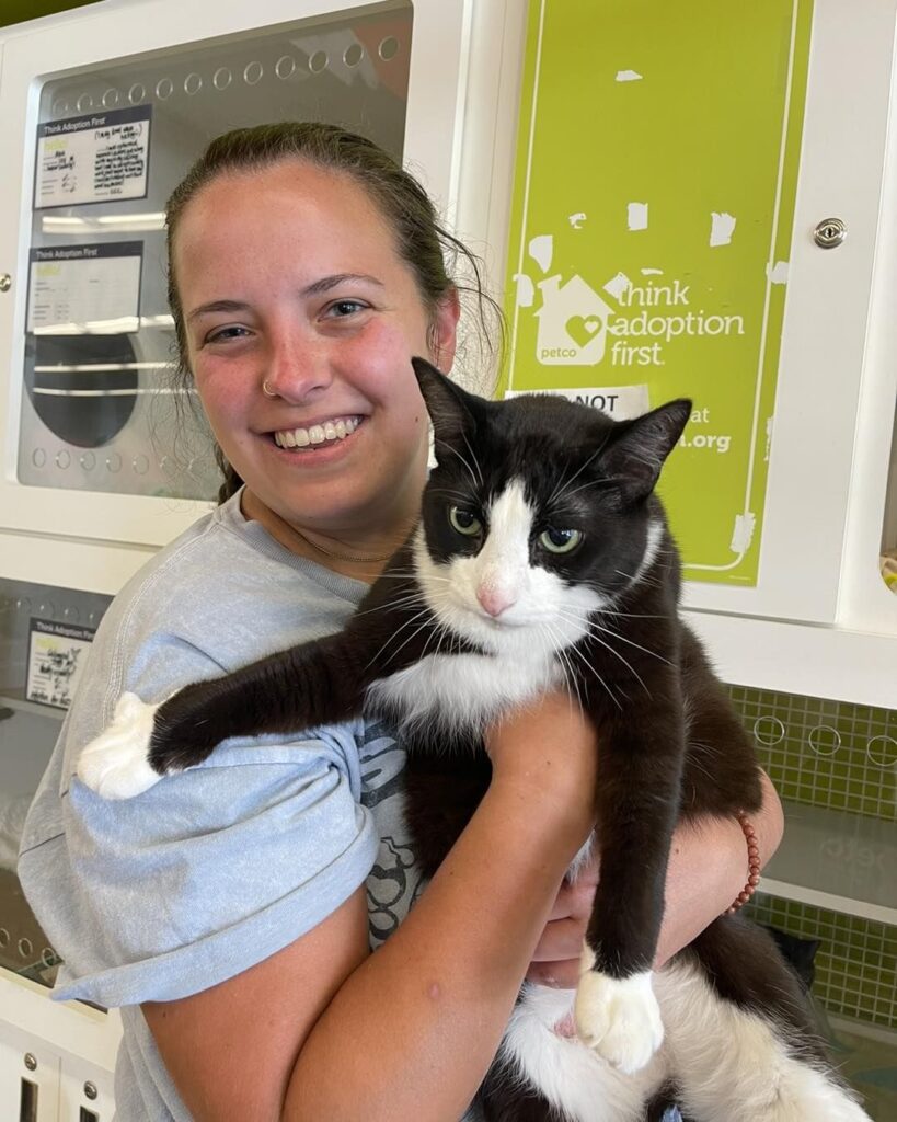 Vali, a black-and-white cat with a permanent toothy grin, finally finds her forever home after two failed adoptions, bringing joy and purrs to her new family.