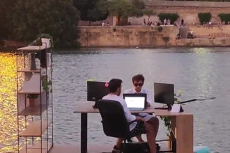 Two lads meet work deadlines on a 'floating office' in Seville, Spain, cruising the Guadalquivir River on surfboards with desks, plants, and computers. TikTok video goes viral.