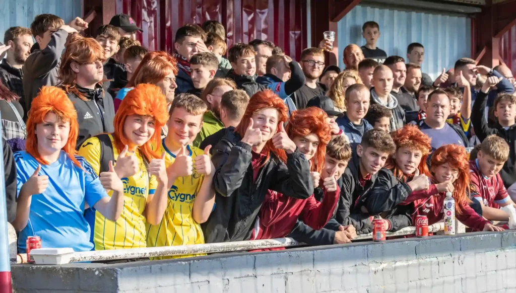 Taunton Town FC fans sport ginger mullet wigs in support of captain Tom Smith, sparking a booming trend at matches. The quirky craze has taken the club's home games by storm!