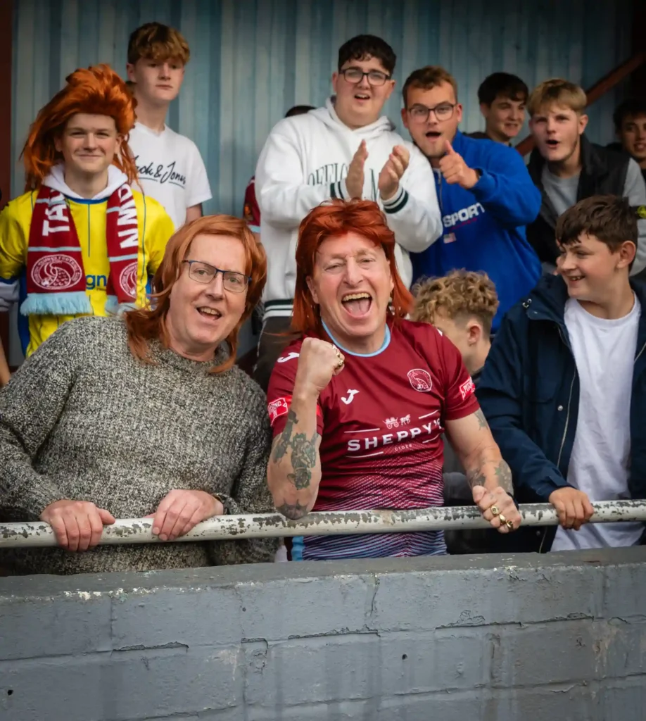 Taunton Town FC fans sport ginger mullet wigs in support of captain Tom Smith, sparking a booming trend at matches. The quirky craze has taken the club's home games by storm!