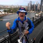 Barmy Army trumpeter Simon Finch sparks Ashes excitement atop Sydney Harbour Bridge, marking one year until England vs Australia 2025 series. Plan your trip now!