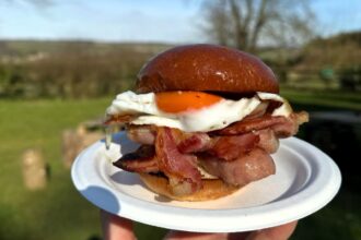 Jeremy Clarkson is facing backlash for charging £7.50 for a bacon roll and £14 for a burger without chips at his country pub, with critics calling the prices greedy.