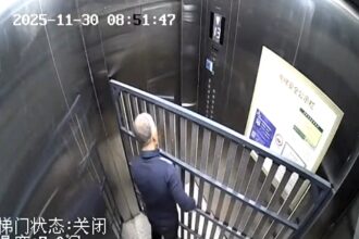 CCTV captures an elderly man stuck in a lift with a bulky bed frame, trapped for 14 minutes as doors jam shut in a slapstick mishap that sent viewers laughing online now