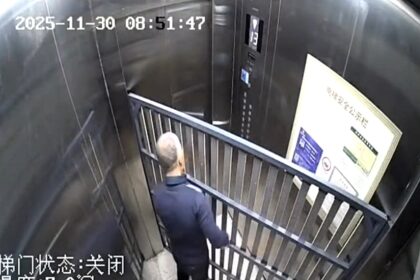 CCTV captures an elderly man stuck in a lift with a bulky bed frame, trapped for 14 minutes as doors jam shut in a slapstick mishap that sent viewers laughing online now