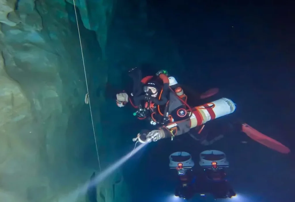 Diver in red and black gear descending through illuminated cave shaft.