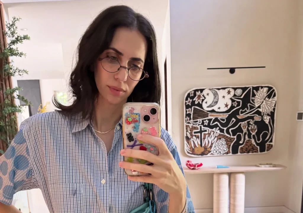 Hila Klein in a striped shirt taking a mirror selfie.