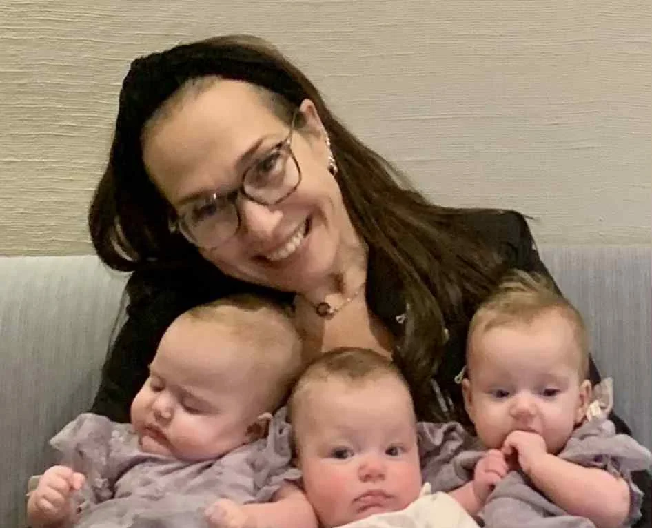 Woman holding three infants in white clothing indoors.
