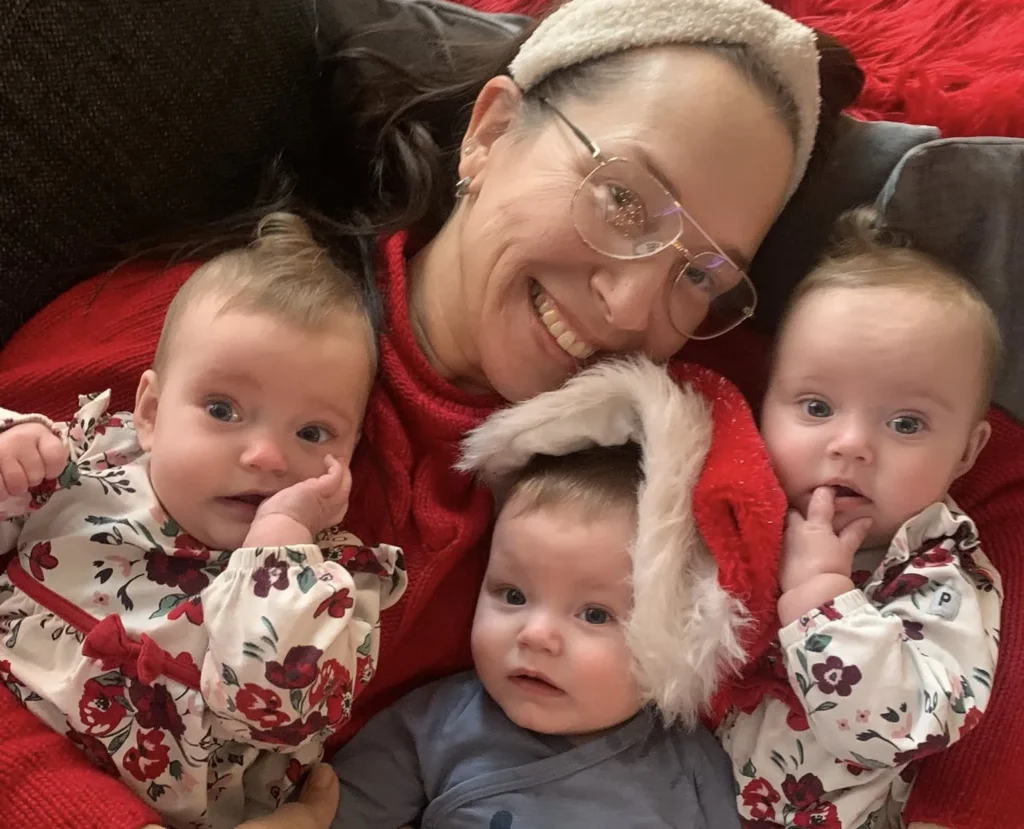Woman in red top holding three infants in patterned clothing on red blanket.