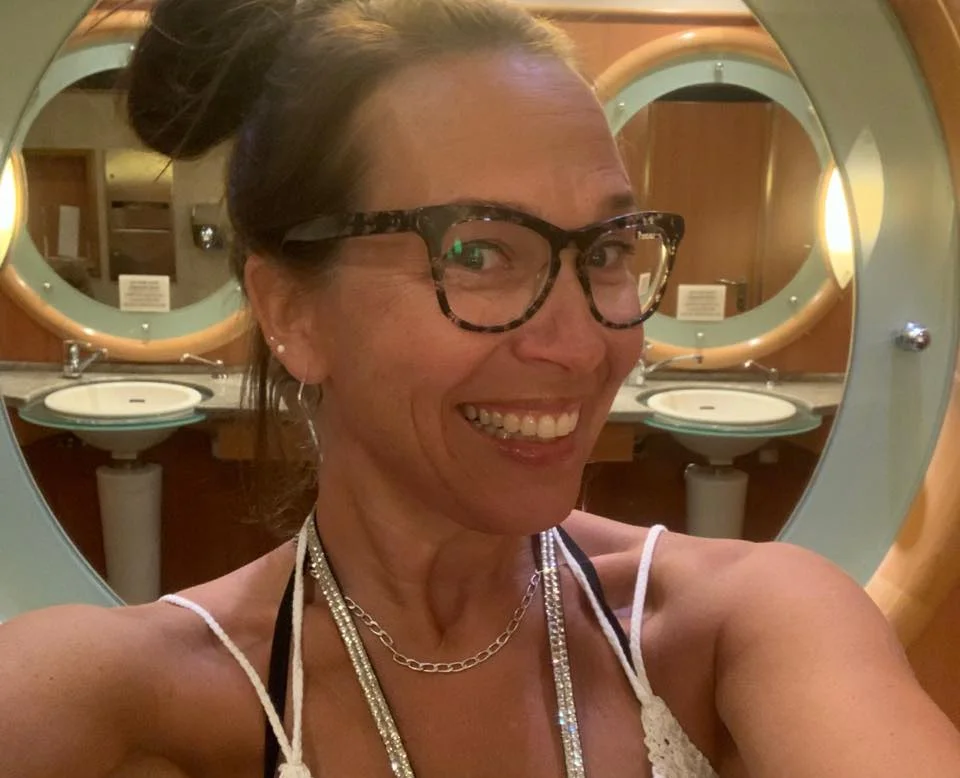 Woman wearing glasses and necklace smiling at camera in bathroom mirror.