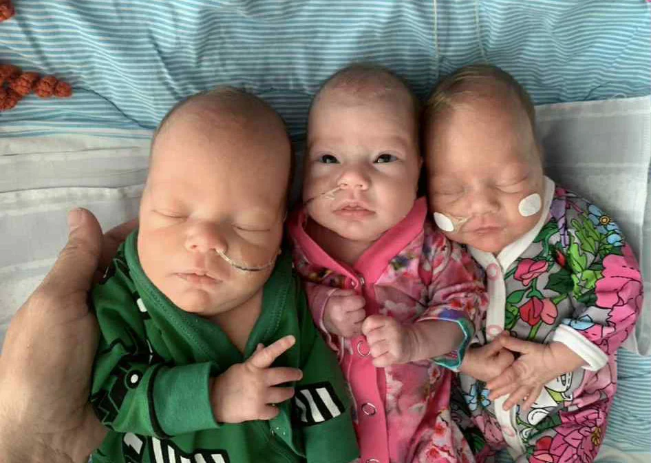 Three newborn babies in patterned clothing lying together on blanket.