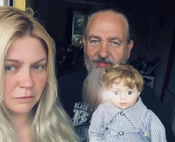 Blonde woman, bearded man and small doll wearing striped shirt posed together indoors