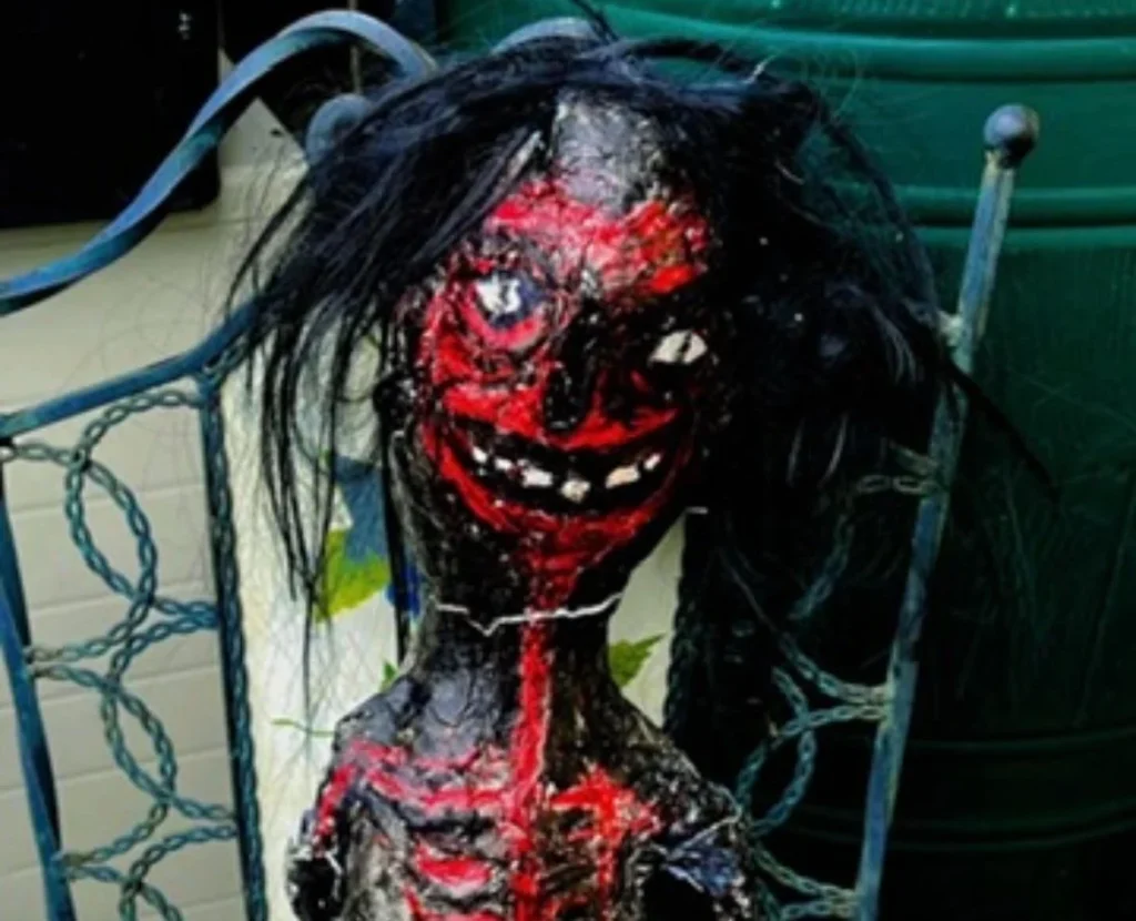 Grotesque doll covered in red paint and black hair, mounted on chair with disturbing expression