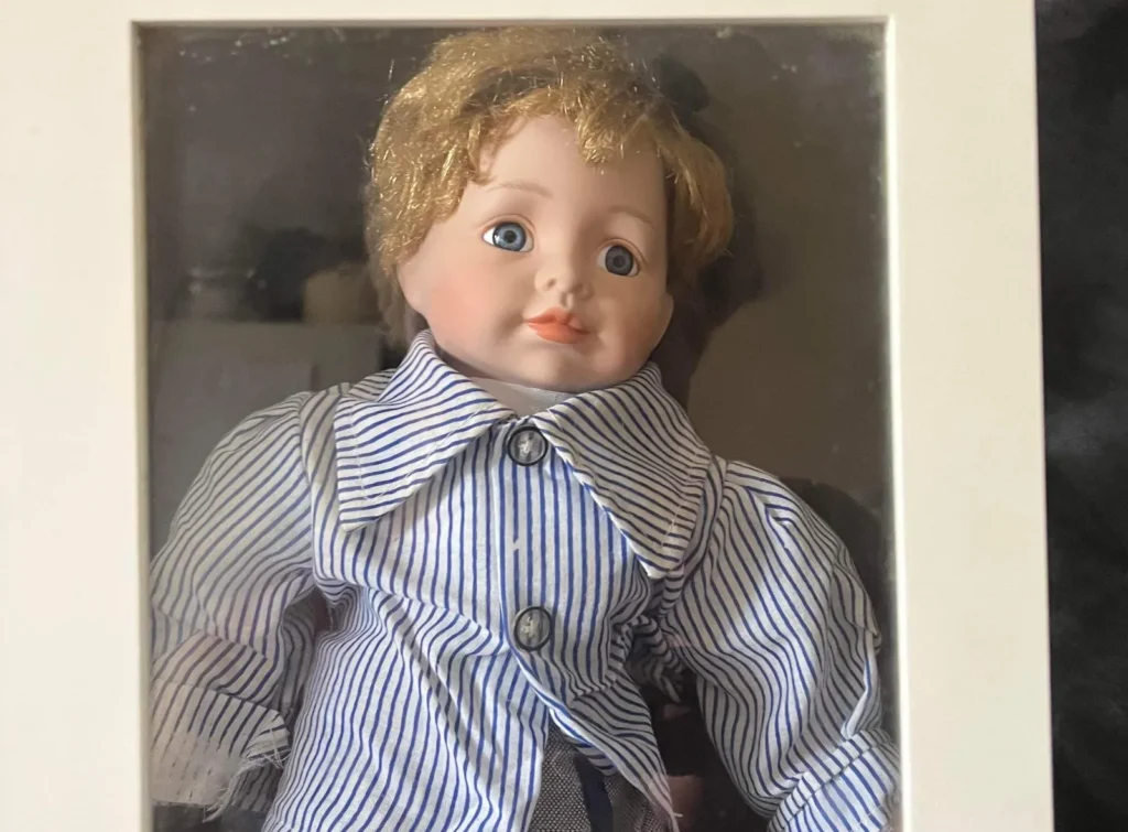 Doll with brown curly hair wearing blue and white striped shirt and grey skirt in box