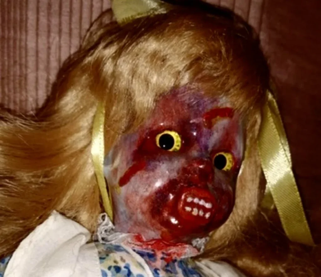 Doll with blonde hair wearing white pinafore with blood-stained face and exposed tendons